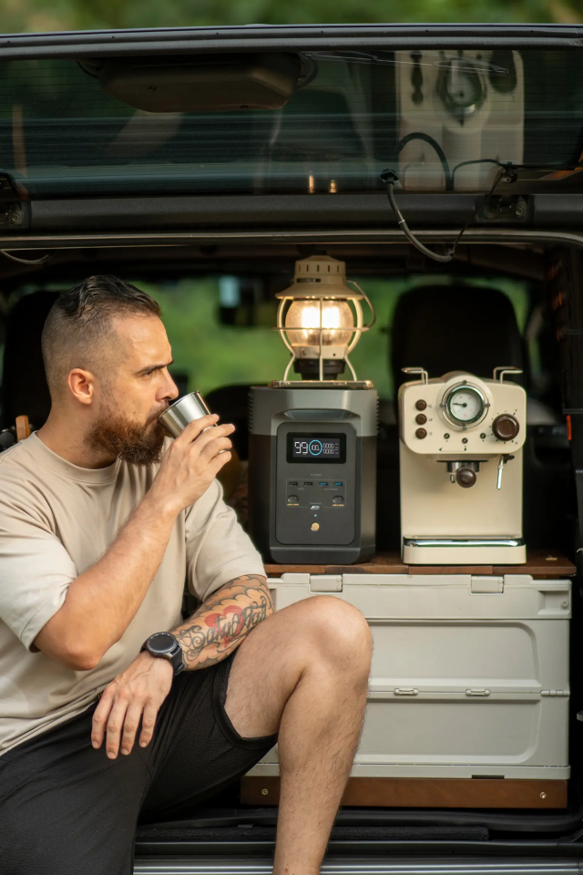 Ein Mann sitzt im Camper und genießt einen Becher Kaffee. Daneben eine Powerstation und eine Retro-Kaffeemaschine.