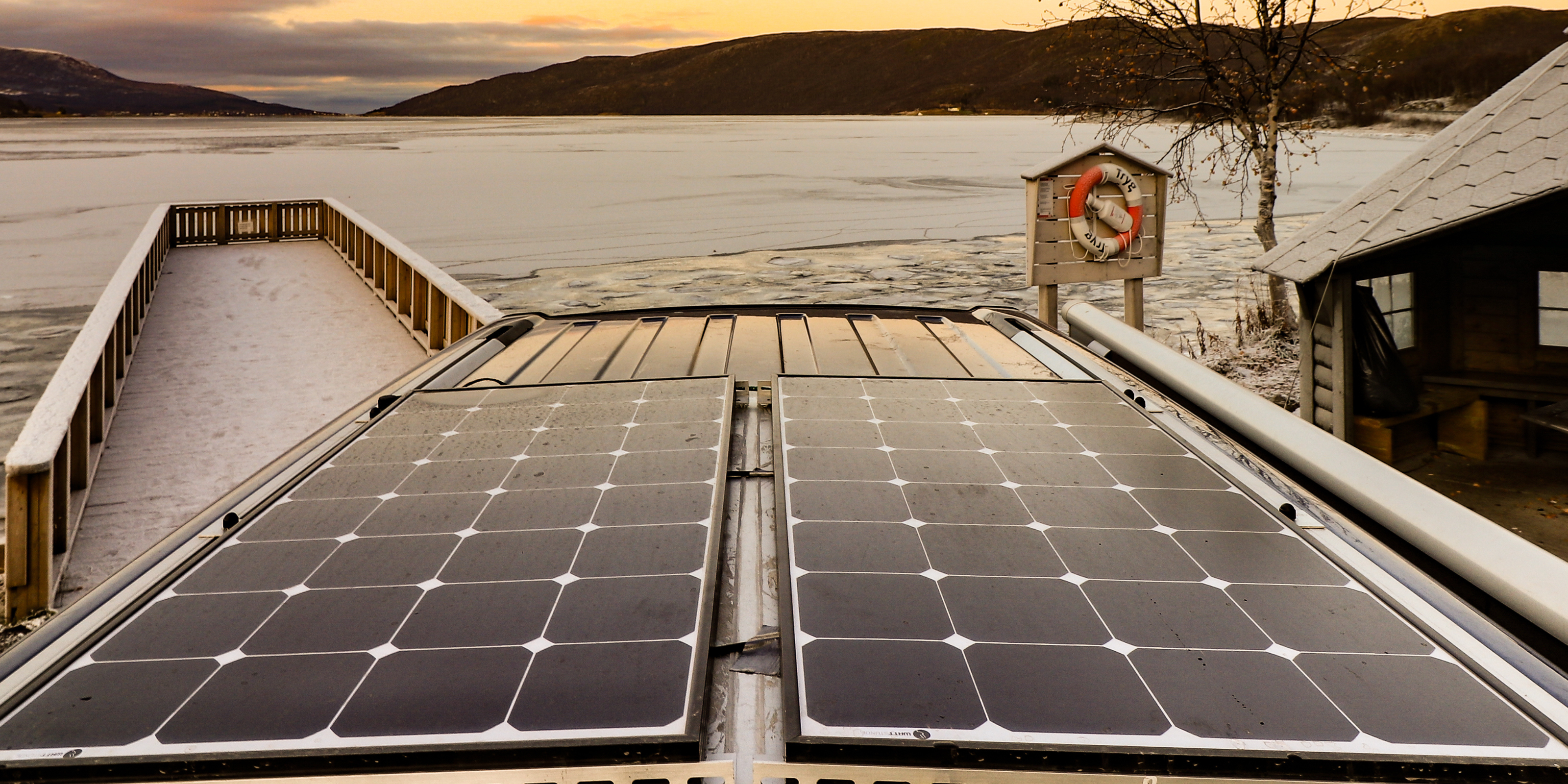 Zwei Solarmodule, die auf dem Dach eines Campers montiert sind. Der Camper selbst steht am See-Ufer , dahinter geht die Sonne unter.
