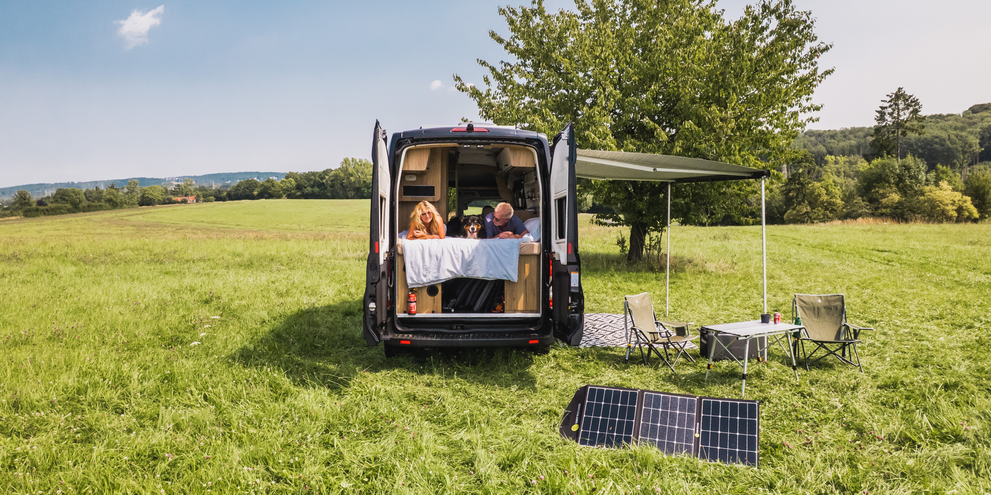 Ein Camper steht mitten in der Natur, davor steht eine Solartasche von WATTSTUNDE. Ein Ehe paar liegt mit ihrem Hund im Bett und sie schauen aus dem Camper heraus.