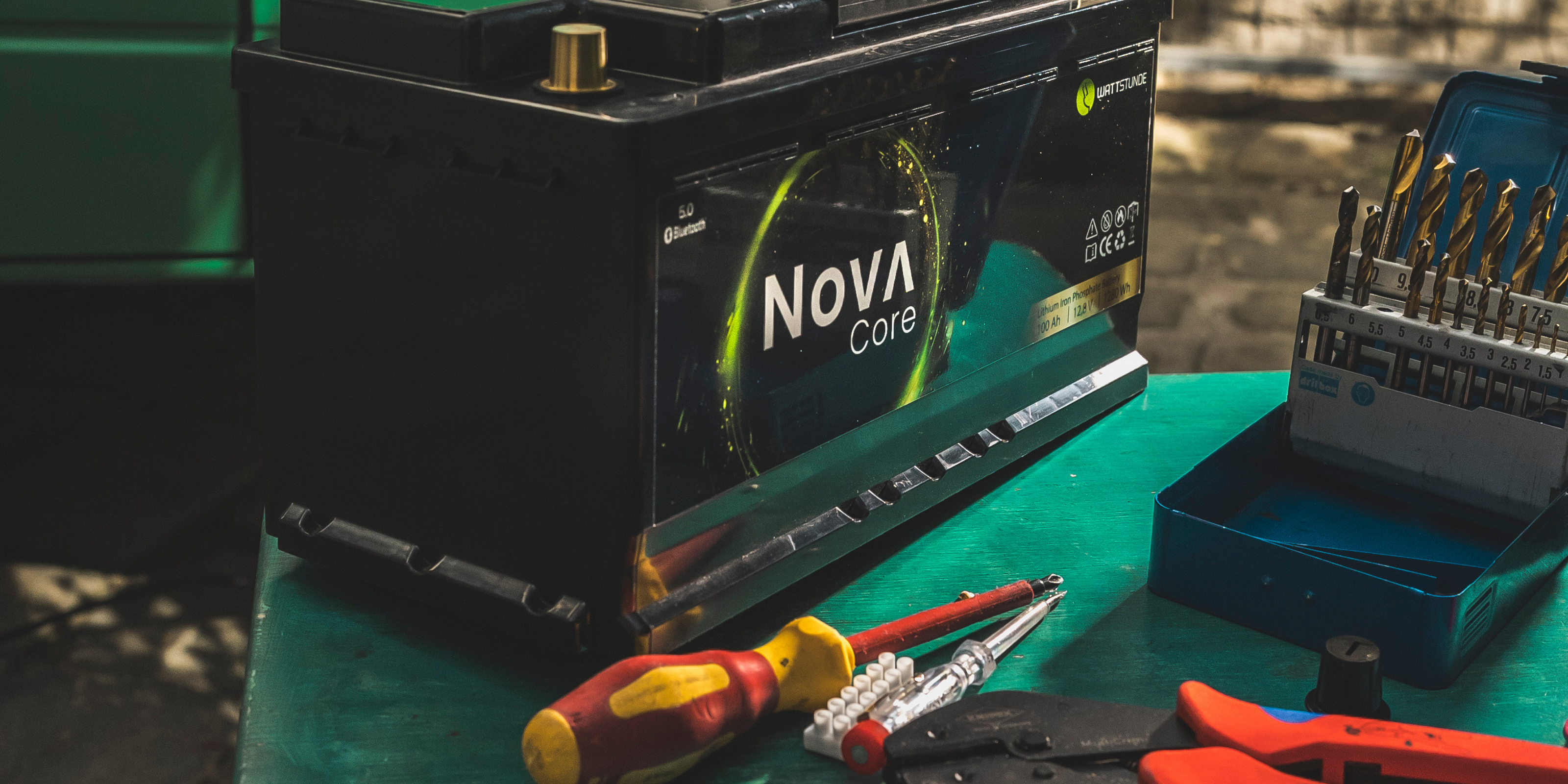 Eine NOVA Core WATTSTUNDE Lithiumbatterie steht auf einem Tisch vor einem Camper. Vor der Batterie liegt Werkzeug.