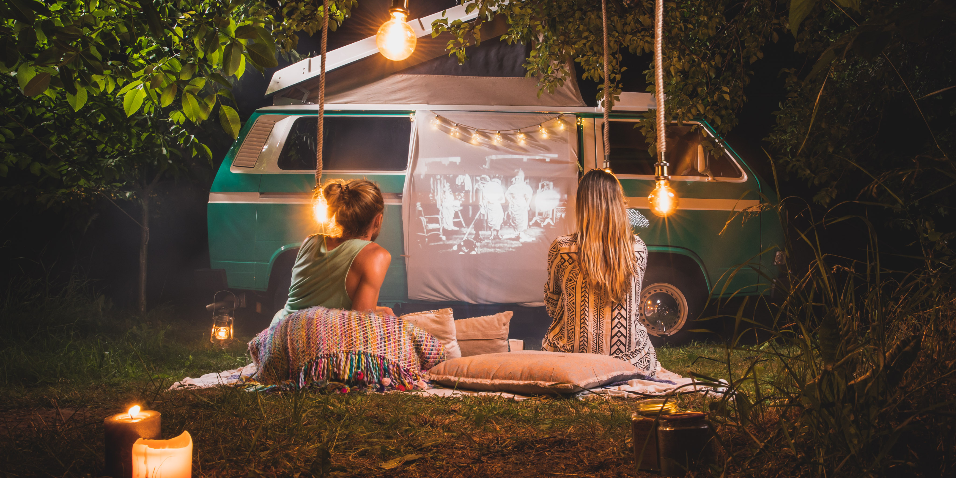 Ein Pärchen sitzt nachts vor dem Wohnmobil in einer romantischen Kulisse mit Lichterketten und schaut dabei einen Film.