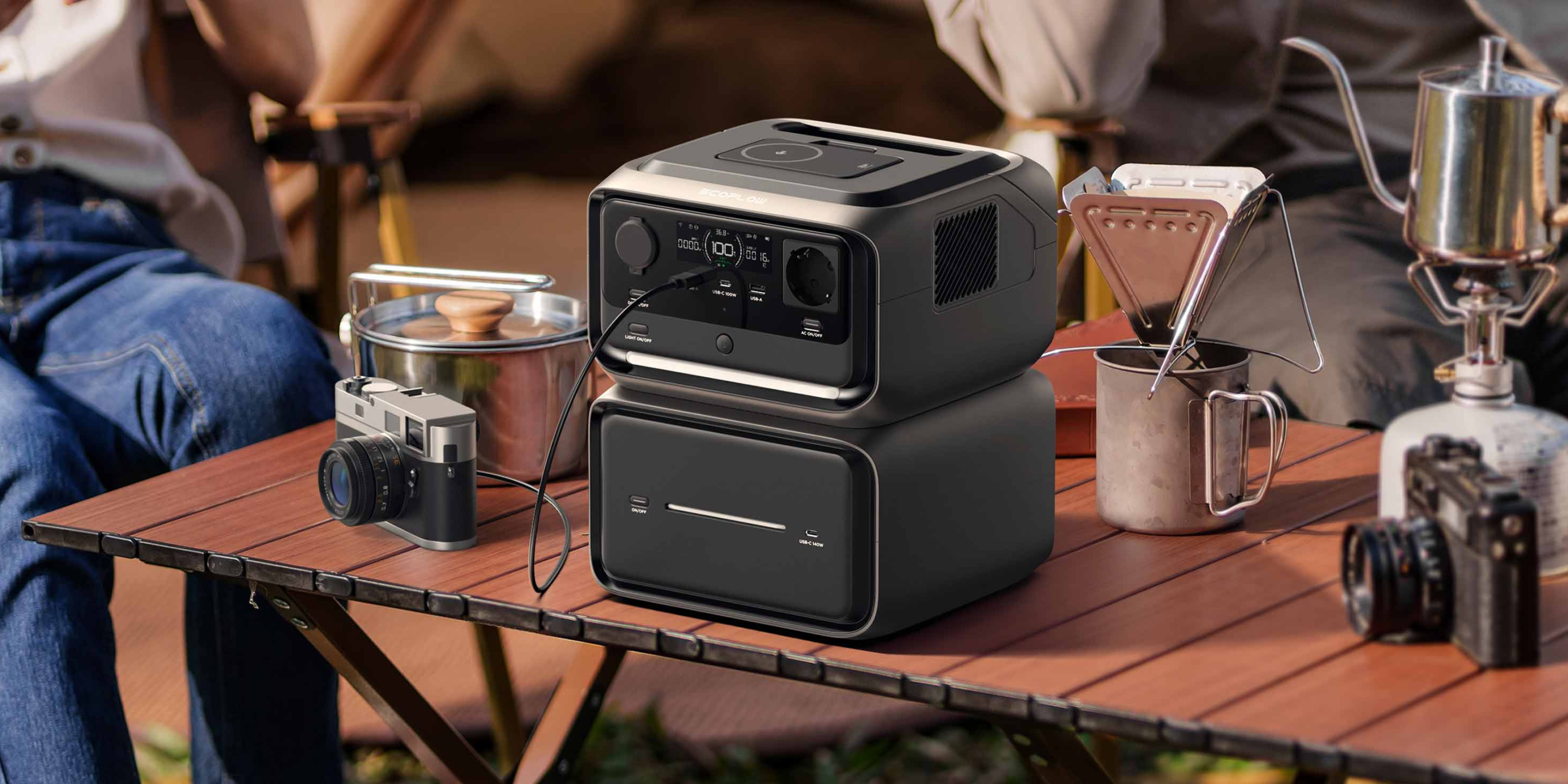 Ein Campingtisch in Holzoptik. Darauf eine Powerstation mit Zusatzbatterie. Daneben sind zwei Kameras, eine davon wird aufgeladenKaffeeutensilien